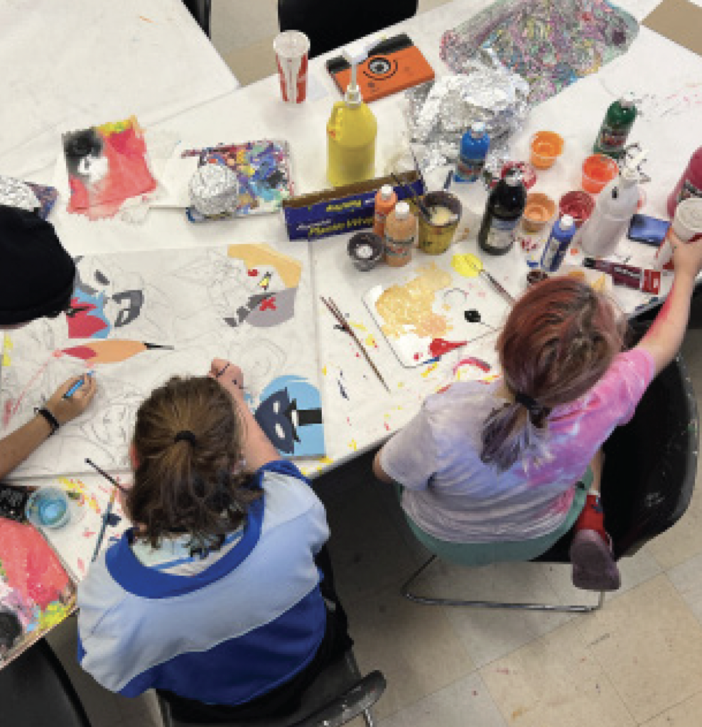 Two people sitting at a table are painting bird-themed artwork. Various paints, brushes, and art supplies are scattered on the table.